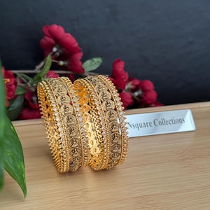 May include: Two gold-colored bangles with intricate patterns and small, clear stones. The bangles are displayed on a wooden surface with red flowers in the background. The text "Nsquare Collections" is visible.