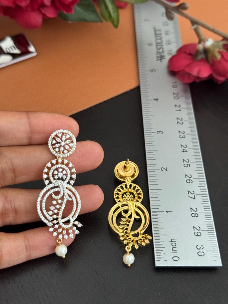 Pu&ograve; includere: Eleganti orecchini dorati e con strass, con un design a tre livelli. Gli orecchini presentano un motivo floreale e una perla pendente. Il righello misura fino a 7.6 cm.