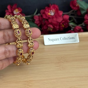 May include: Two gold-colored bangles with intricate designs and small red gemstones. The bangles feature detailed figures and oval-shaped red stones. The background includes red flowers and a wooden surface. The text "Nsquare Collections" is visible.