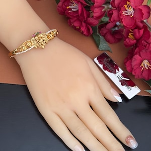 Op de afbeelding: Goudkleurige armband met ingewikkelde details, met een centraal ontwerp met rode en groene accenten. De armband is versierd met kleine, heldere stenen. De afbeelding toont ook een hand met een French manicure en een kaart met de tekst "Nsquare Collections".