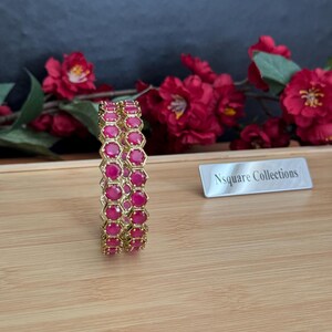 May include: Two gold bangles with a honeycomb design, each cell filled with a vibrant pink gemstone. The bangles are displayed on a wooden surface, with red flowers in the background. The text "Nsquare Collections" is visible.