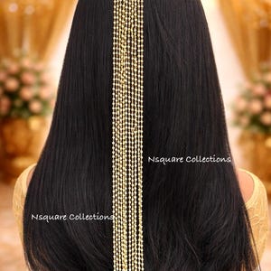 Peut inclure: Un accessoire de cheveux doré avec un dessus décoratif orné de pierres précieuses rouges, vertes et transparentes. De longues mèches de petites perles rondes descendent dans le dos. Le texte "Nsquare Collections" est visible.