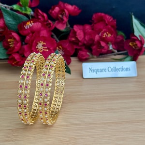 May include: Two gold bangles featuring a central row of alternating round pink and white stones, framed by delicate scalloped edges. A small rectangular sign with the text Nsquare Collections is visible in the background.