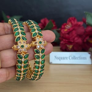 Peut inclure: Deux bracelets dorés ornés de pierres vertes en forme de feuilles et d'accents en cristal transparent. Les bracelets présentent un motif floral au niveau du fermoir. Le texte "Nsquare Collections" est visible sur une petite étiquette blanche.