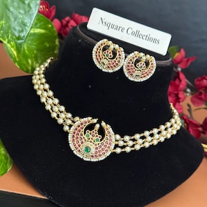 May include: A gold-toned necklace and earring set featuring three strands of small white pearls. The necklace and matching circular earrings display a crescent-shaped pendant adorned with red and green stones. A white tag reads Nsquare Collections.