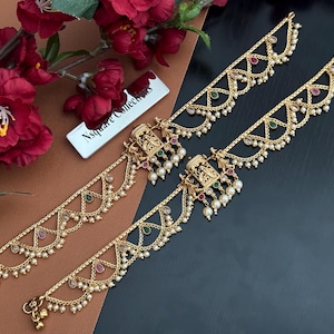 May include: Gold-toned hair jewelry with intricate designs, featuring small pearls and colorful gemstones. The pieces are arranged on a brown and black surface, with a sprig of red flowers in the background. The jewelry has a traditional Indian aesthetic.