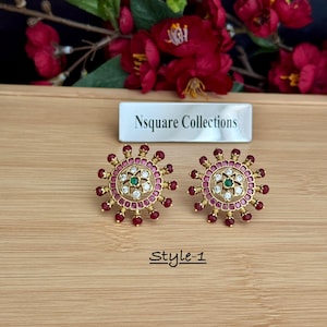 May include: A pair of gold-toned stud earrings. Each earring features a central green stone surrounded by small white stones and a ring of red beads. The earrings are on a wooden surface, with the text "Style-1" below.