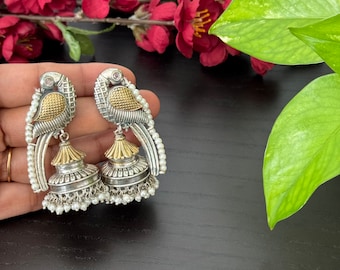 May include: A pair of silver and gold earrings with a bird design and pearl accents. The earrings are hanging from a hand.