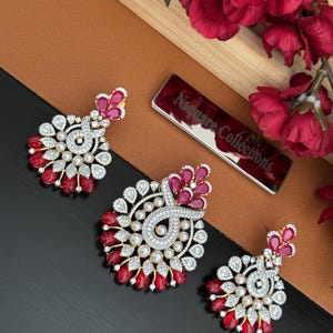 May include: Three ornate, gold-toned earrings with a floral design. Each earring features a cluster of red gemstones, white pearls, and clear crystals. The earrings are displayed on a brown and black surface, with a red floral arrangement in the background.