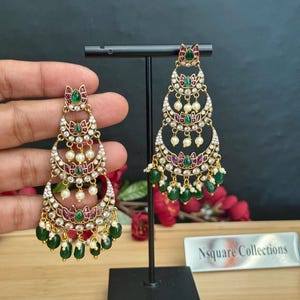 May include: Gold-toned chandelier earrings with a multi-tiered design. Each tier features a crescent shape adorned with clear crystals, small pearls, and ruby-colored and emerald-green accents. The earrings have a dangling bead design.