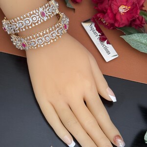 Peut inclure: Deux bracelets dorés avec des motifs ajourés complexes, ornés de pierres transparentes et rouge rubis. Les bracelets sont présentés sur une main de mannequin avec une manucure française. Le fond comprend des fleurs rouges et une étiquette argentée avec le texte "Nsquare Collections."