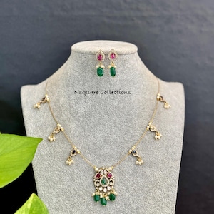May include: A gold-toned necklace and matching earrings set. The necklace features a central pendant with red, green, and white gemstones, and dangling green beads. The earrings have red and green gemstones. The text "Nsquare Collections" is visible.