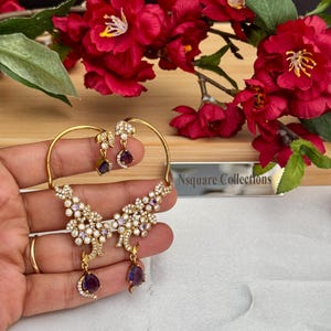 May include: Gold-toned floral earrings with dangling purple teardrop-shaped gems. The earrings feature a cluster of small white stones and are held in a hand. The background includes red flowers and the text "Nsquare Collections".