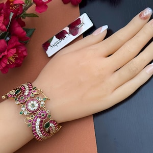 May include: A gold-toned bracelet featuring intricate peacock-inspired motifs adorned with vibrant red and green stones, along with clear white accents. The design includes circular and curved elements, showcasing a detailed metalwork pattern.