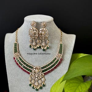 May include: An ornate gold-toned necklace and earring set. The necklace features green and red gemstones, with a central pendant and dangling accents. The earrings are circular with dangling accents. The text "Nsquare Collections" is visible.
