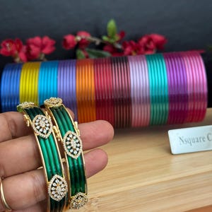 May include: Two emerald green bangles featuring gold-tone metal accents and clear crystal embellishments in a marquise-shaped pattern. Behind them, a large collection of thin, metallic-finish bangles in a rainbow of colors is displayed.