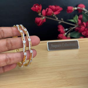 Peut inclure: Deux bracelets joncs dorés présentant un design ondulé incrusté de petites pierres transparentes. Les bracelets ont une finition polie et un motif répétitif de grappes de pierres circulaires le long de la surface ondulée des anneaux.