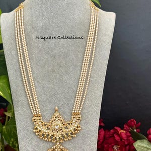May include: A multi-strand necklace featuring four rows of small white beads connected to an ornate gold-tone pendant with clear stones and green teardrop accents. The text Nsquare Collections is printed on the gray display bust.