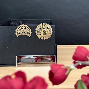 Könnte beinhalten: Ein Paar goldfarbene Ohrringe mit einem halbmondförmigen Design und einem kreisförmigen Design mit einem zentralen Pfauenmotiv. Beide Ohrringe sind mit klaren Steinen sowie kleinen grünen und roten Akzenten verziert und liegen auf einer schwarzen Box.