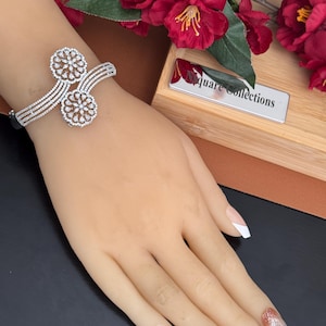 May include: A silver-toned cuff bracelet featuring four parallel bands that converge into two circular floral motifs. Each circular element is encrusted with numerous small, clear, sparkling stones arranged in a symmetrical, petal-like pattern.