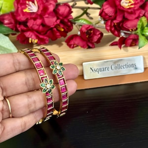Może przedstawiać: Dwie złote bransoletki z czerwonymi prostokątnymi kamieniami i zielonymi kwiatowymi akcentami. Bransoletki trzymane są w dłoni, z widocznym złotym pierścionkiem. Tło przedstawia czerwone kwiaty i drewnianą powierzchnię.