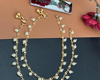 Può includere: Un paio di cavigliere color oro con piccole perle pendenti. Le cavigliere sono esposte su una superficie marrone, con uno sfondo sfocato che include una composizione floreale e un biglietto da visita.