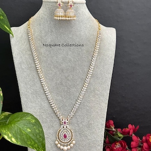May include: A long necklace and matching jhumka earrings set featuring clear white stones in a leaf-like pattern. The necklace has a central teardrop-shaped pendant with a pink stone and small white pearls. The text Nsquare Collections is visible.