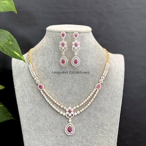 May include: A gold-toned necklace and earring set featuring teardrop-shaped pink stones surrounded by clear crystals. The necklace has a double-strand design with a central floral motif and a dangling teardrop pendant. Text reads Nsquare Collections.