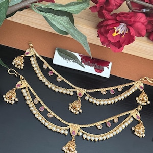May include: Gold-toned ear chains with three strands of faux pearls and red accents. Each chain features a bell-shaped charm and is designed to be worn with earrings. The chains are displayed on a dark surface with floral accents.