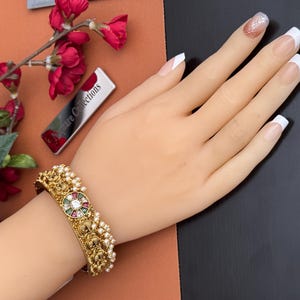 May include: A gold-toned bracelet with a central circular design featuring a white gem surrounded by red, green, and gold accents. The bracelet is adorned with small pearls and intricate carvings. The hand is on a brown and black surface.