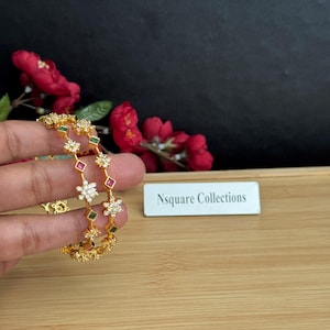 May include: Gold-toned bangles with floral and geometric accents. The bangles feature clear and colored gemstones, including white, green, and red. The text "Nsquare Collections" is visible on a white label.