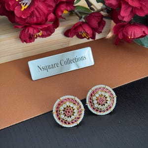 以下が含まれることがあります： 金色の縁取りが施された2つの丸いスタッドピアス。中央には赤と透明な宝石のデザインが施されています。ピアスは小さな白い真珠で囲まれています。「Nsquare Collections」の文字が銀色のネームプレートにあります。