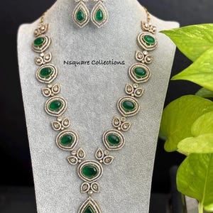 Może przedstawiać: Złoty naszyjnik z długim, łzowym wisiorem i owalnymi zielonymi kamieniami osadzonymi w srebrnej ramie. Naszyjnik zdobią mniejsze białe kamienie. W zestawie znajdują się również pasujące kolczyki z zielonymi i białymi kamieniami. W obrazie widoczny jest tekst "Nsquare Collections".