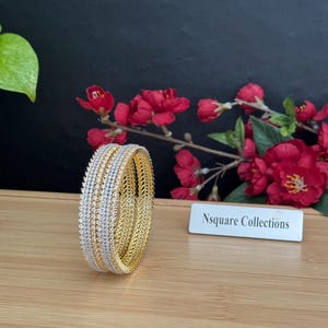 May include: Gold and silver bangles with a sparkling design. The bangles are stacked together, showcasing a mix of gold and silver tones. The background features red flowers and the text "Nsquare Collections".