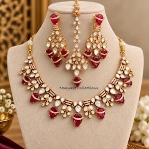 May include: A gold-toned jewelry set featuring a necklace, two drop earrings, and a maang tikka. Each piece is adorned with deep red teardrop-shaped stones and clear white crystals. The text Nsquare Collections is visible on the necklace.