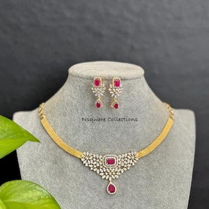 May include: A gold-toned necklace and earring set with red and clear gemstones. The necklace features a central rectangular red stone with a teardrop pendant. The earrings have matching rectangular red stones. The text "Nsquare Collections" is visible.