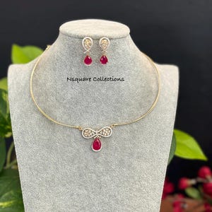 May include: A gold-toned necklace and earring set displayed on a gray mannequin. The necklace features a pendant with a red teardrop-shaped stone. The earrings also have red teardrop stones. The text "Nsquare Collections" is visible.