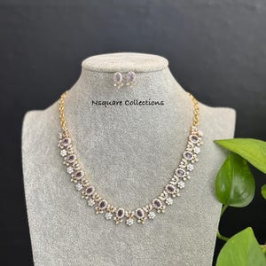 Puede incluir: Un conjunto de collar y pendientes a juego en tono dorado. El collar presenta piedras ovaladas moradas alternas y pequeñas piedras blancas. Los pendientes tienen un diseño similar. El texto "Nsquare Collections" es visible.