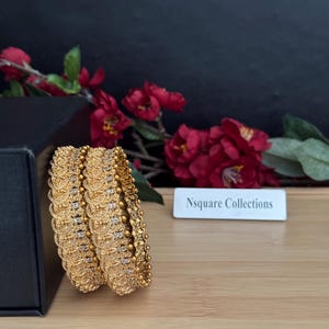 Puede incluir: Brazaletes dorados con intrincados diseños y pequeños detalles brillantes. Los brazaletes están apilados, mostrando su diseño detallado. El fondo incluye flores rojas y un letrero que dice "Nsquare Collections."