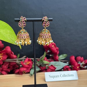 May include: Gold-toned, ornate earrings with a peacock design. The earrings feature red and green gemstones, with a bell-shaped bottom adorned with small white pearls. The earrings are displayed on a black stand against a dark background.