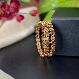 Può includere: Due braccialetti d'oro con disegni intricati e pietre preziose quadrate rosse. I braccialetti sono su una superficie nera.