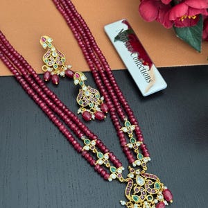 May include: A maroon beaded necklace and matching earrings set. The necklace features three strands of small beads, with gold-colored pendants and accents. The earrings have a similar design. The set is embellished with green and red gemstones. The tag reads "Nsquare Collections."