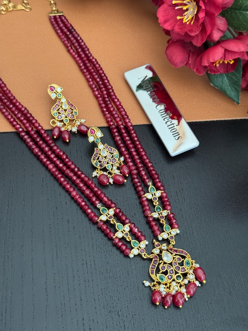 May include: A maroon beaded necklace and matching earrings set. The necklace features three strands of small beads, with gold-colored pendants and accents. The earrings have a similar design. The set is embellished with green and red gemstones. The tag reads "Nsquare Collections."