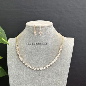 May include: A gold-toned necklace and matching drop earrings set featuring a repeating pattern of clear, baguette-cut and marquise-cut stones. The jewelry set is displayed on a gray bust, with the text Nsquare Collections printed in black.