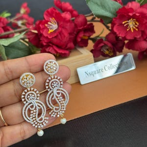 Pu&ograve; includere: Eleganti orecchini color oro con intricati disegni traforati, incastonati con scintillanti cristalli trasparenti. Ogni orecchino presenta una piccola perla pendente nella parte inferiore. Gli orecchini sono esposti su uno sfondo di fiori rossi.