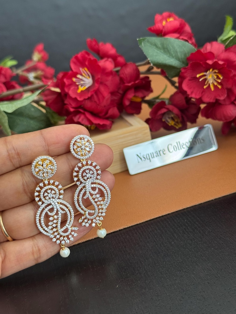 Pu&ograve; includere: Eleganti orecchini color oro con intricati disegni traforati, incastonati con scintillanti cristalli trasparenti. Ogni orecchino presenta una piccola perla pendente nella parte inferiore. Gli orecchini sono esposti su uno sfondo di fiori rossi.
