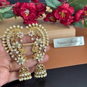 May include: A pair of gold earrings with a peacock design. The earrings have detailed patterns, small white pearls, clear and coloured gemstones, and a bell-shaped base. They are shown against a backdrop of red flowers.