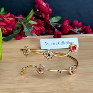 May include: Two gold-colored bangles with floral designs and colorful gemstones. The bangles are adorned with clear and red gemstones, and a green gemstone in the center of a flower. The text "Nsquare Collections" is visible.