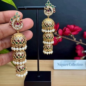 Può includere: Orecchini pendenti dorati con dettagli intricati. Ogni orecchino presenta una serie di gocce ornate a forma di campana adornate con pietre preziose rosse, verdi e trasparenti e piccole perle bianche. Gli orecchini sono esposti su un supporto nero.