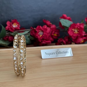 Op de afbeelding: Twee gouden armbanden met heldere edelstenen, gepresenteerd op een houten dienblad met rode bloemen op de achtergrond. De armbanden hebben een gedetailleerd ontwerp met vierkante en ovale stenen. Een klein wit bordje met de tekst "Nsquare Collections" is ook zichtbaar.